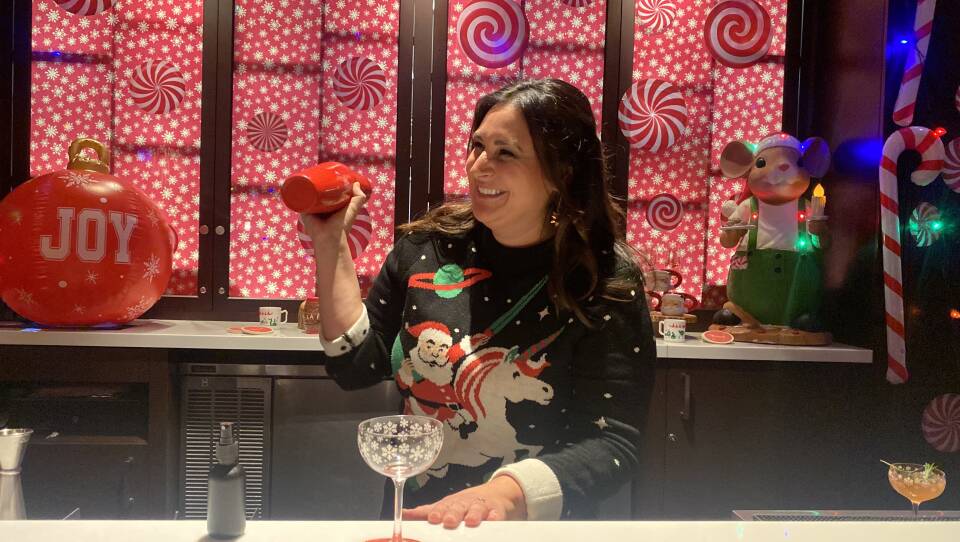 A woman in a sweater showing Santa riding a unicorn stands behind a bar, shaking a cocktail shaker.