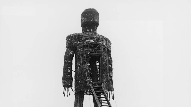 A black and white photo of a giant statue of a man, perhaps 30 feet tall. Four actors stand around it with tiki torches.