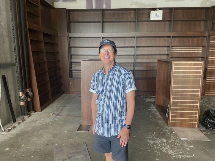 This is a photograph of Jeff Peters who is opening up a new bookstore. In this photograph he is stnading in the bookstore wich is under construction. The room is full of empty bookcases. Jeff Peters is wearing a strpied t-shirt, shorts and a blue baseball cap with an anchor embroidered on the front. He is smiling.