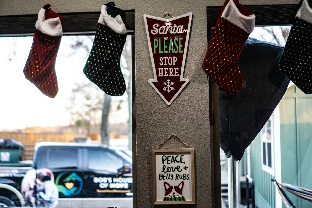 Four stockings hang above windows that face a parking area. A sign shaped like a downward arrow reads "Santa please stop here"