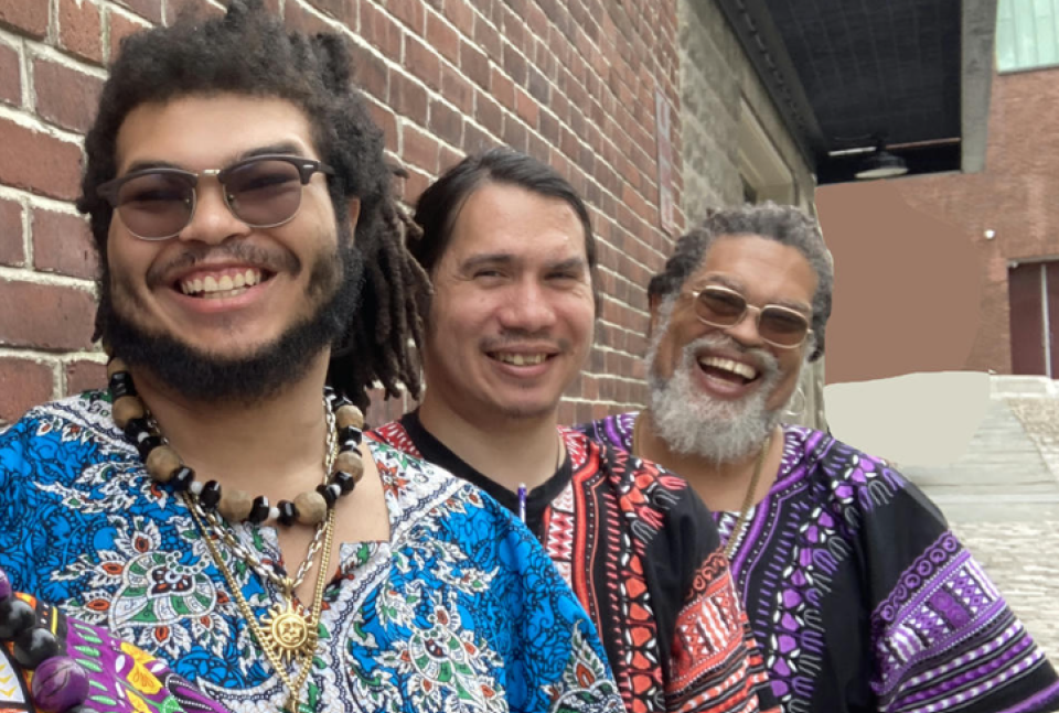 This is a photo of the Grammy-nominated soul-funk-blues band The GroovaLottos. In this image three of the band members are standing outdoors, against a brick wall. They are wearing brightly colored dashiki shirts. They are looking at the camera and smiling--big smiles.