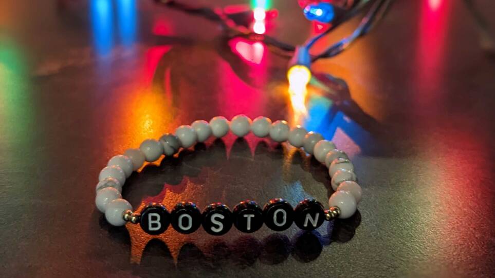A beaded bracelet on a black table with Christmas lights behind it. The bracelet has black beads with white letters spelling out the word Boston.