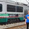 A woman in a shirt reading Metro Housing Boston stands in front of train tracks. Behind her, a Green Line trolley passes on the tracks. A Packard's Corner station sign is visible.