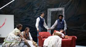 In this scene from the opera La Boheme,  Mimi has succumbed to her long illness. he body  lies on an orange, velvet fainting couch. On the floor, to her left, her lover Rodolfo has collapsed on the floor in despair