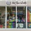 This is a photograph of a storefront of a book store. Hanging in the window is a multi colored striped Progress Pride flag, there is also a Pride Flag. in the storefront window is a table, which has  a display of books propped on it and a Pride flag draped over it. Propped up in the window sill is a Black Lives Matter poster with the face of George Floyd , a painting of a red heart and a sign in the window, which says "All She Wrote Books. Celebrate Trans Writers & Stories Every Day"