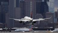A passenger plane with its wheels lowered comes down to land at an airport while other planes taxi on the ground nearby. Boston's downtown skyline is in the background.