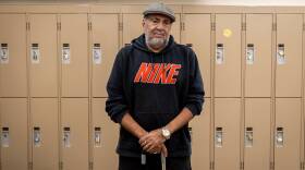 A man in a black hoodie and a grey cap stands in front of beige lockers holding a cane.