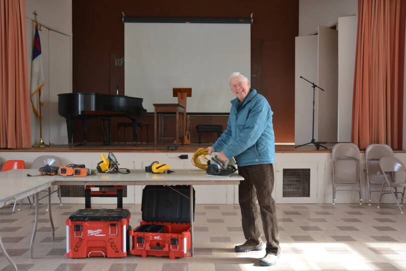 An older white man wearing a blue jacket and brown pants, smiles with teeth, as he works with tools on a table. He stand in a former church auditorium, it has beige tiled floors, a stage with a podium and grand piano are in the background.