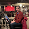 A woman smiles at the camera. She is sitting in the back of a theater venue. She has a music stand with an iPad and iPhone in front of her and a microphone.