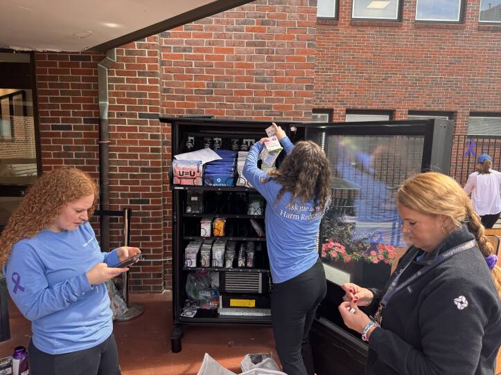 Three people are standing about a vending machine, stocking it with public health items outside of a brick hospital.