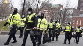 a group of state troopers walks in boston, wearing warm black clothes like hats and layers under bright yellow vests, carrying sticks, helmets and other gear while wearing masks