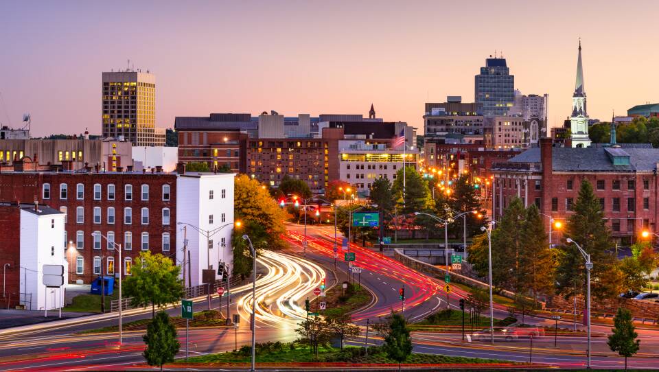 The headlights and taillights of cars light up the roadway into a city's downtown area with plenty of large buildings and a speckling of trees.