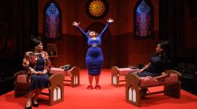 A woman in a blue dress stands in front of rows of pews with her arms raised. Two women seated in the pews look at her.