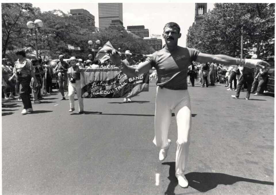 A black and white photo of a man waving a rainbow flag, in front of a parade going down a main city street