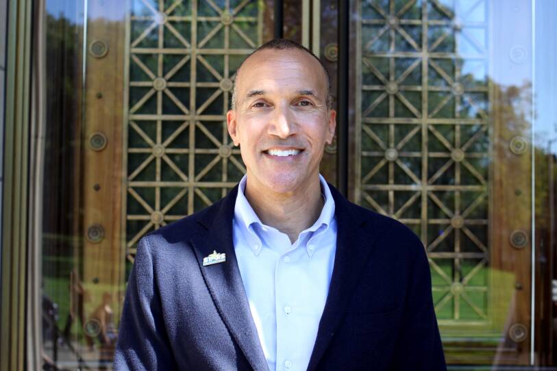 A man in a blazer poses, smiling, in front of an academic building.
