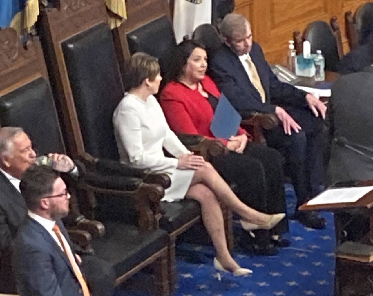 A woman sits in a large, ornate wood chair. Her feet don't touch the carpet.