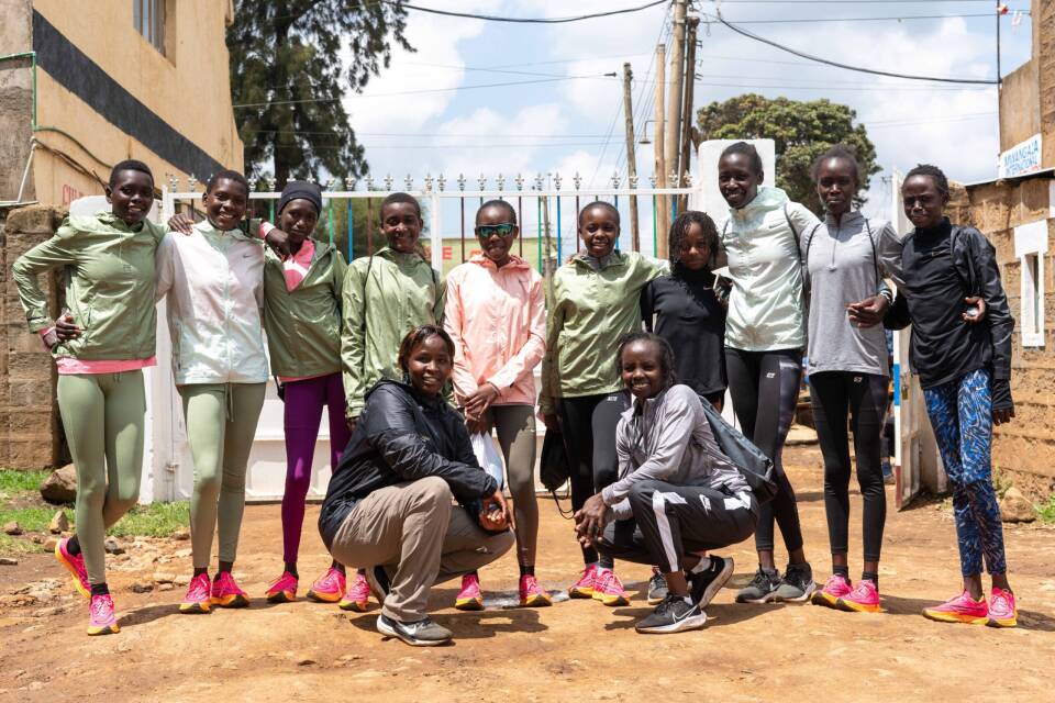 A dozen girls in athletic clothing stand on the dirt road in a village.