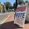 A white voting sign with 'VOTE' and an American flag sits at an street intersection with a yellow-leaved tree in the background