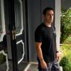 A man in a T shirt stands on the front porch of a home. He looks concerned and is resting his arms at his sides.