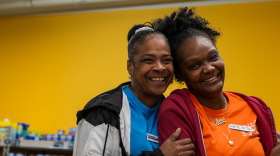 Two women with hoodies and name tags hug and smile for the camera. A grocery store shelf is visible in the background.