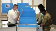 A corrections officer in uniform speaks two inmates wearing olive drab clothing as they stand at a row of blue voting booths.