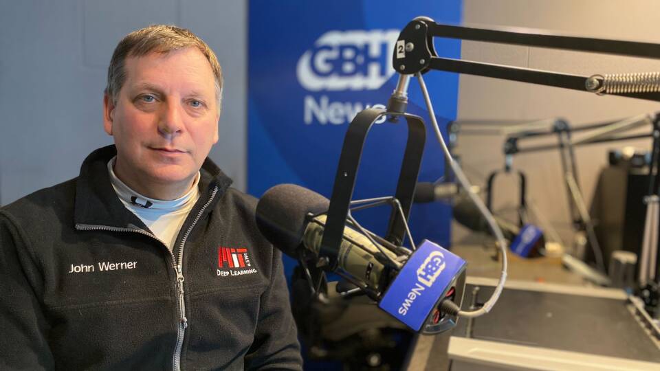 A man in a zip-up fleece with the name John Werner and MIT Deep Learning logo on it sits next to a microphone in a radio studio.