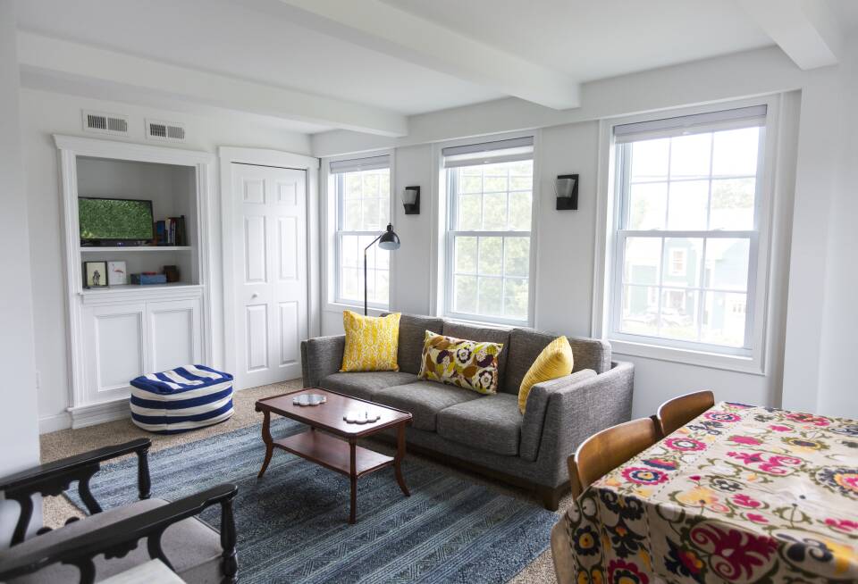 A cozy living room with a gray couch, wood coffee table and brightly accented pillows and a tablecloth on the dining table to the side.