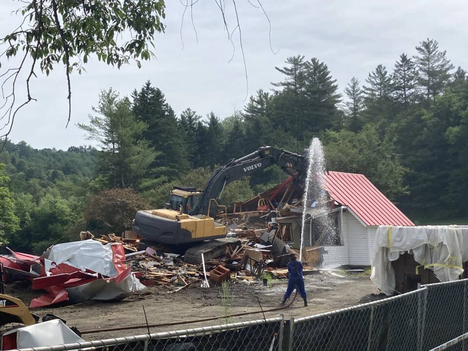 An excavator tears down a building while a person sprays a fire hose.