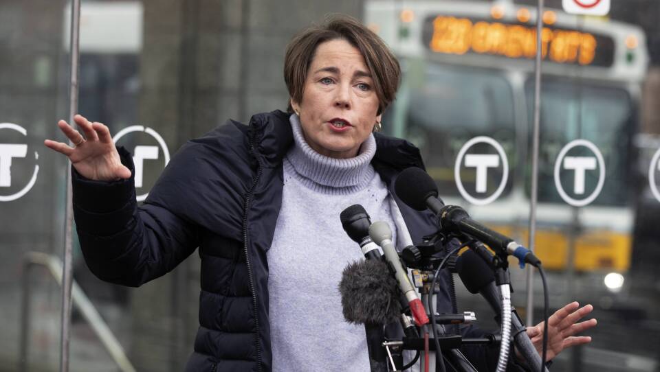 A woman with short brown hair is mid-speech in front of a cluster of microphones, wearing a light purple turtleneck under a winter coat with the reflection of an MBTA bus in the background.