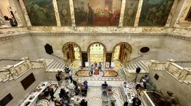 A spacious, two-story marble lobby is the setting for a handful of people sitting in chairs and three figures at a podium beneath arched doorways and large murals.