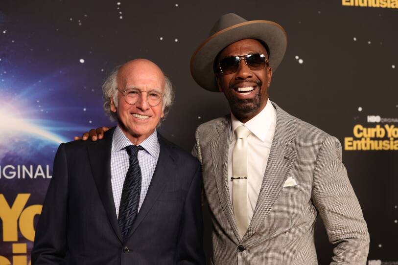 Two men in suits smile for the cameras, Larry David wearing wire-rimmed glasses and Smoove wearing sunglasses with a suede gray hat