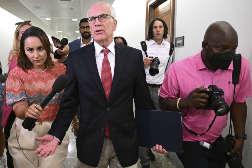 A man in a black blazer and red tie stands facing the camera with his hands outstretched and a folder in one arm. On his right side is a reporter with a microphone and on his left is a photographer with a camera.