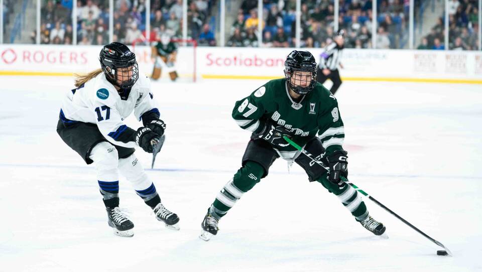 Two women skating, the Boston player handling the puck. Crowds can be seen packed in the stands.
