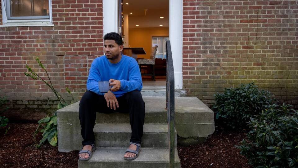 A man in a blue shirt sits on a front stoop, holding a mug.