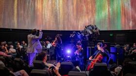 A conductor leads musicians in the center of a small planetarium, surrounded by audience members whose eyes are fixed on the ceiling where bright animated colors fill the screen