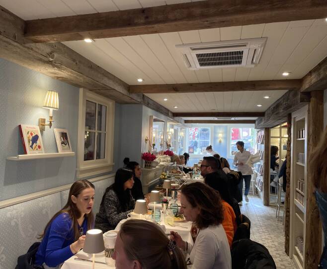 This is an interior photograph of a cafe. People sit along a narrow row of tables with white table cloths. It is a crowded and convivial scene, with people talking. In the background are two servers . Against the back wall are two windows with sunlight streaming through them.