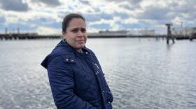 A woman in a blue jacket stands facing the camera in front of a background of water and maritime infrastructure.