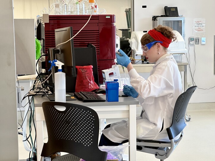 A young white woman with bright red lipstick and a red headband and wearing a white lab coat and blue rubber gloves holds a syringe to work on a lab project.
