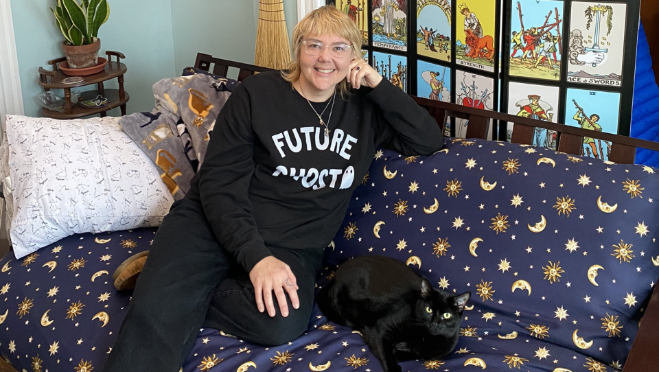 A woman with short blonde hair sits on a futon with a moon, sun, and stars print, a black cat laying next to her. Behind them is a wall of large tarot cards.