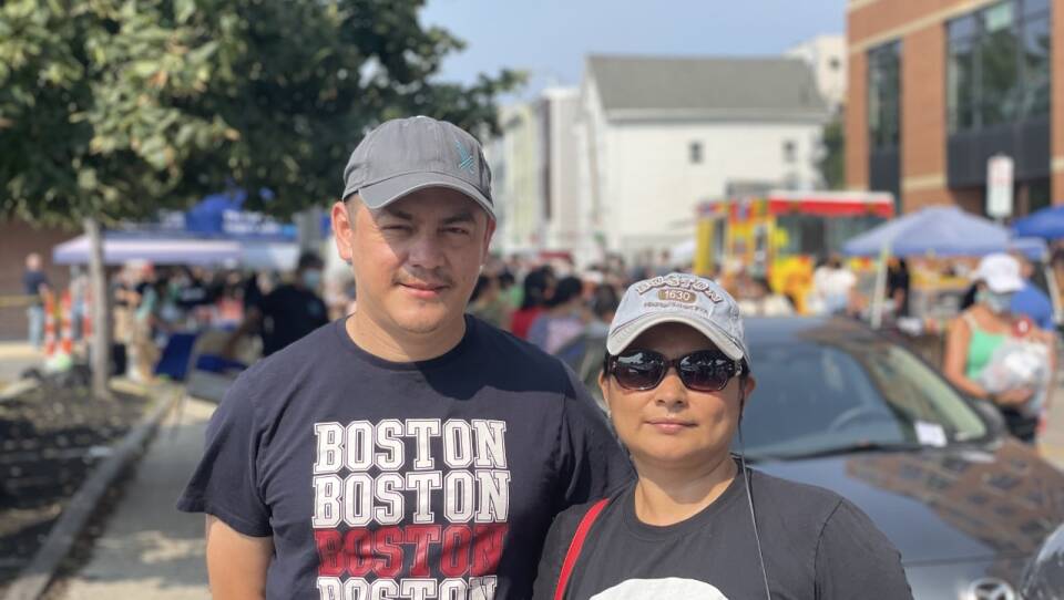 A man and woman wearing baseball caps stand on a street in East Boston.