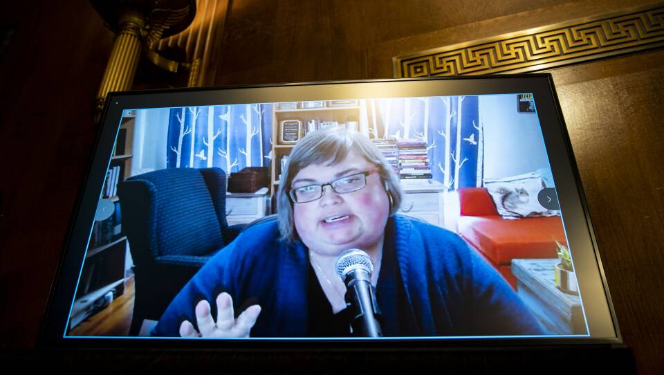 A woman on a video conference app is displayed on a screen in a formal Washington, D.C. hearing room.