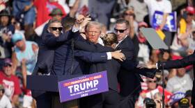 Donald Trump, with a few streaks of blood on his cheek and jaw, is surrounded by agents blocking his body while on stage at a rally.