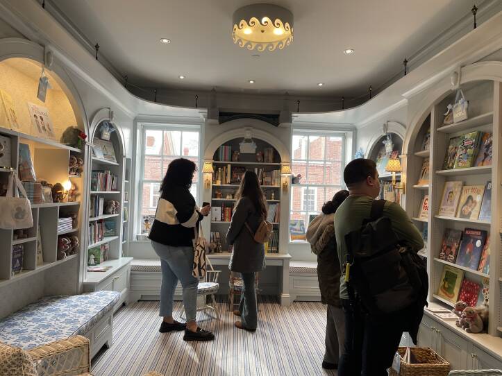 Four people gather in the children's section of a bookstore. The room is designed to look like a child's play room, the colors are light, there are stuffed animals on the bookshelves. The atmosphere is both tranquil and happy.