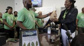 A man behind a counter and register hands a brown paper bag to a woman. Other people stand around watching.
