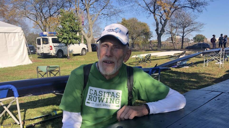 An old, bearded man wearing a Casitas Rowing club shirt sits at a picnic table in the shade.