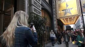 A woman on a city sidewalk takes a photo with her cellphone of the "Hamilton" musical sign outside a theater.