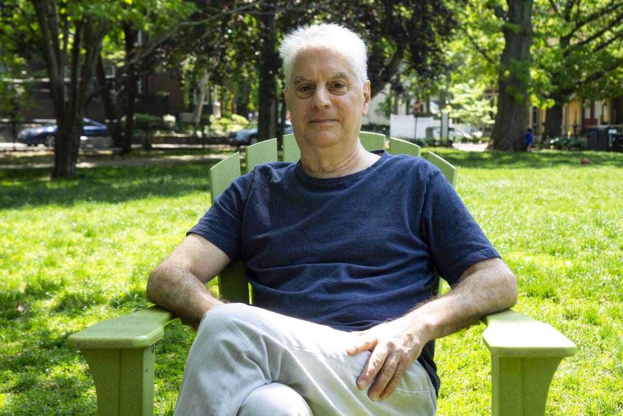 An older man sits in a green Adirondack chair in a grassy park lined with trees.