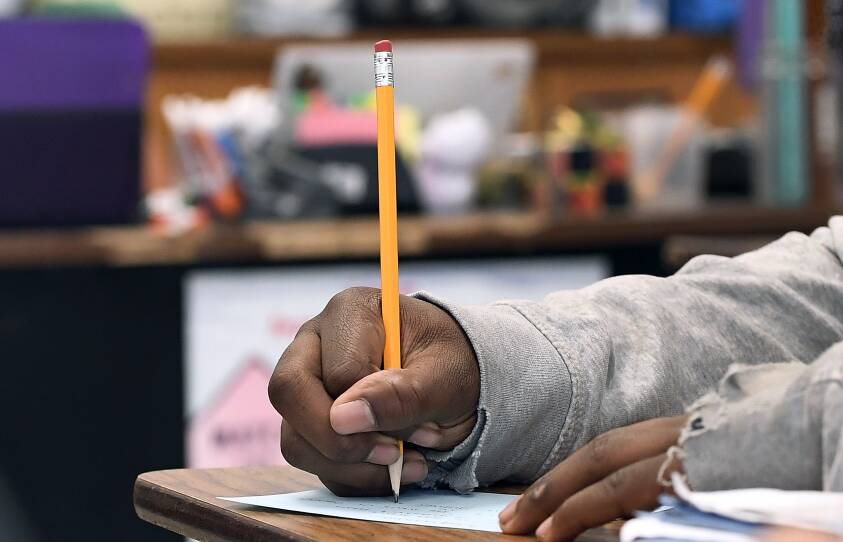 A Black student writes with a pencil at their desk in a classroom