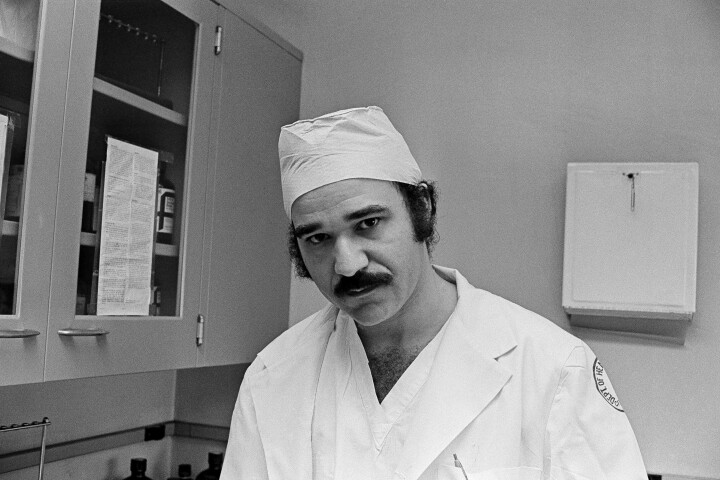 A black and white photograph of a man wearing a doctor's uniform in a hospital exam room.