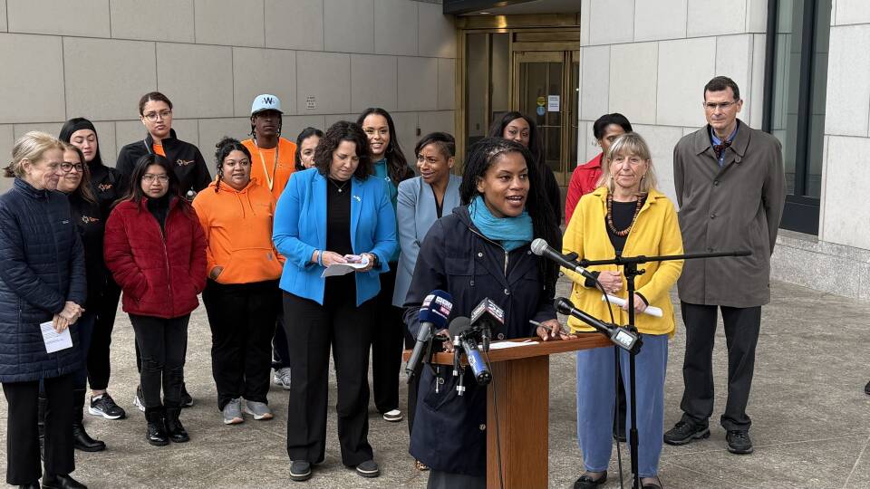 A woman gives remarks from podium while more than a dozen people behind her listen on outside a courthouse building in Boston.